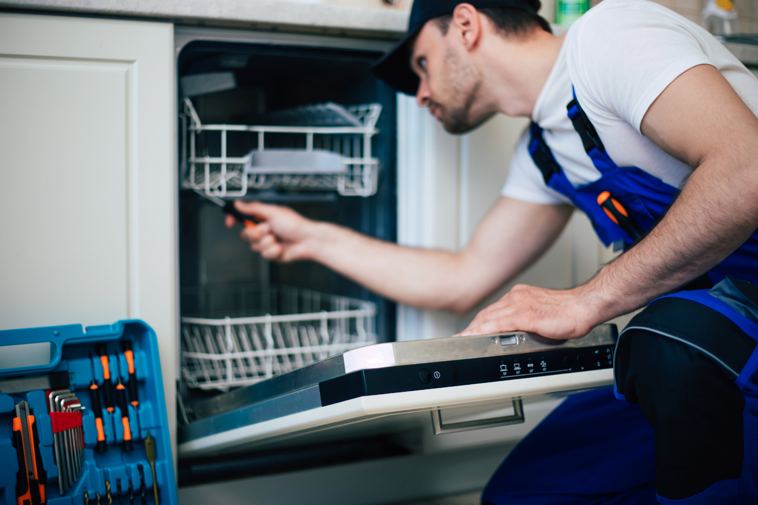 A male technician wearing a black cap and blue overalls kneels in a kitchen, repairing a built-in Bosch dishwasher. He is using a tool to inspect the interior of the unit. A blue open toolkit with various screwdrivers and bits is visible in the foreground.