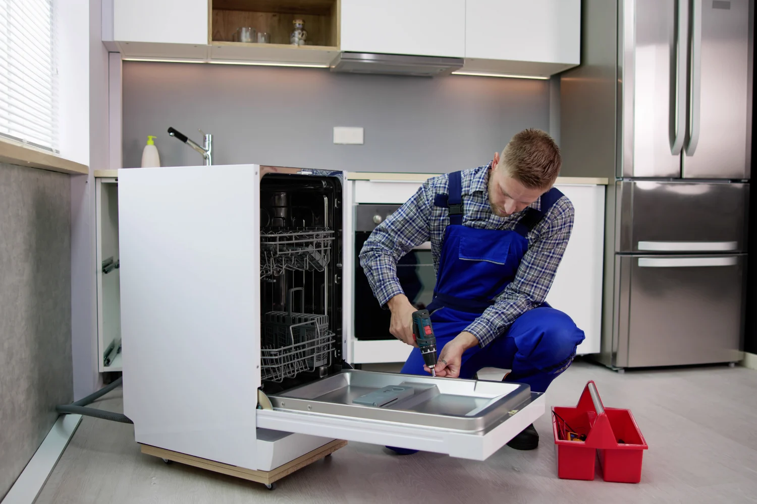 A technician in a plaid shirt and blue overalls kneels on a kitchen floor, using a power drill to work on the open door of a white dishwasher. The dishwasher has been pulled out from the cabinetry, and a red tool caddy sits on the floor nearby.