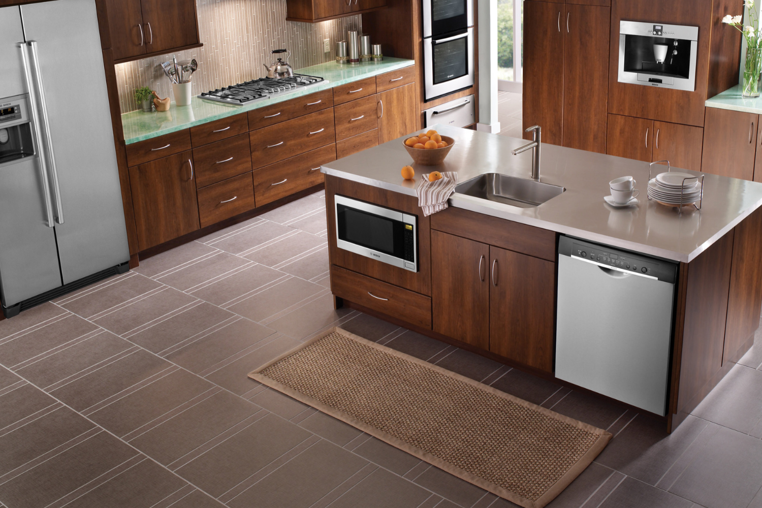 A high-angle view of a spacious kitchen featuring dark wood cabinetry and a full suite of stainless steel Bosch appliances, including a refrigerator, double wall ovens, a built-in coffee maker, a cooktop, and a dishwasher and microwave installed in the central island.