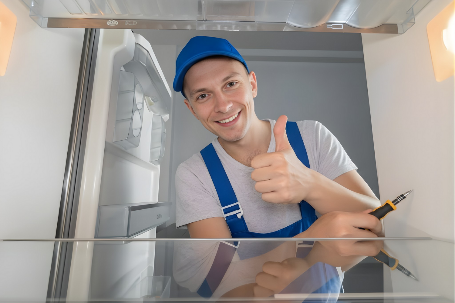 A smiling, factory-certified refrigeration repair technician in a blue uniform gives a thumbs-up from inside an open, clean refrigerator, indicating a successful repair.