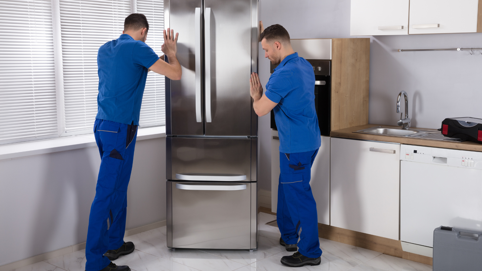 Two service technicians in blue uniforms installing a stainless steel refrigerator in a home kitchen.