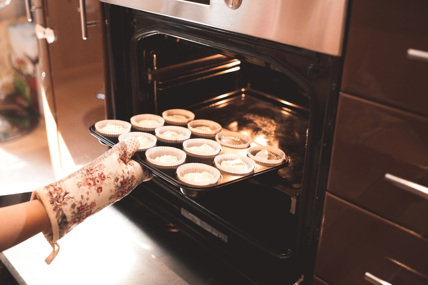 A person wearing a floral patterned oven mitt places a black baking tray filled with raw muffin batter into a warm, lit oven.