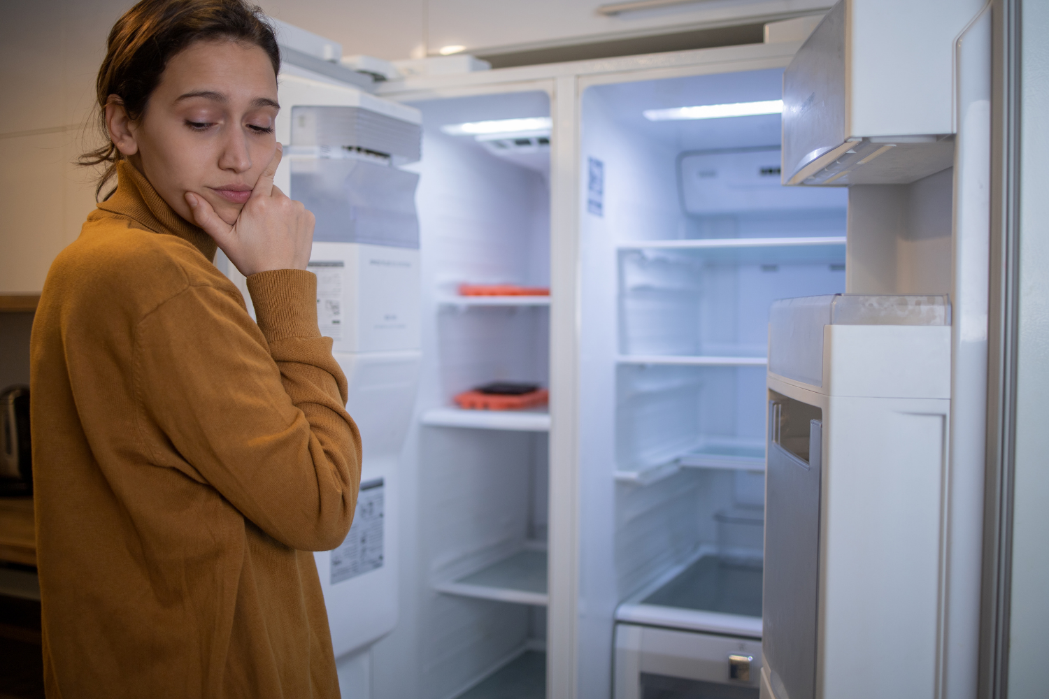 A worried homeowner looks into her empty, open refrigerator, pondering the need for professional refrigeration repair due to cooling failure.