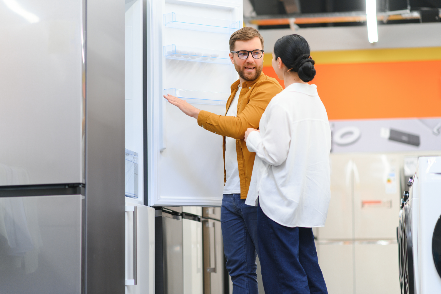 A couple shopping for appliances, with the man wearing glasses pointing inside an open, empty stainless steel refrigerator while discussing it with the woman beside him in a store.
