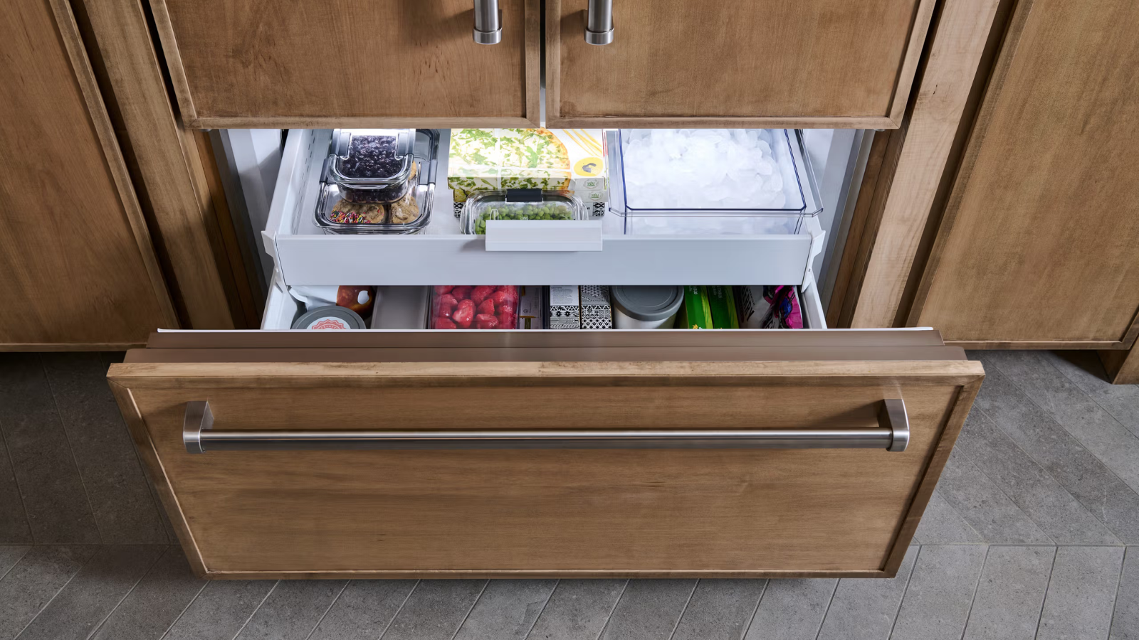 A wooden refrigerator drawer is pulled open to reveal containers of blueberries, salad mix, strawberries, and ice.