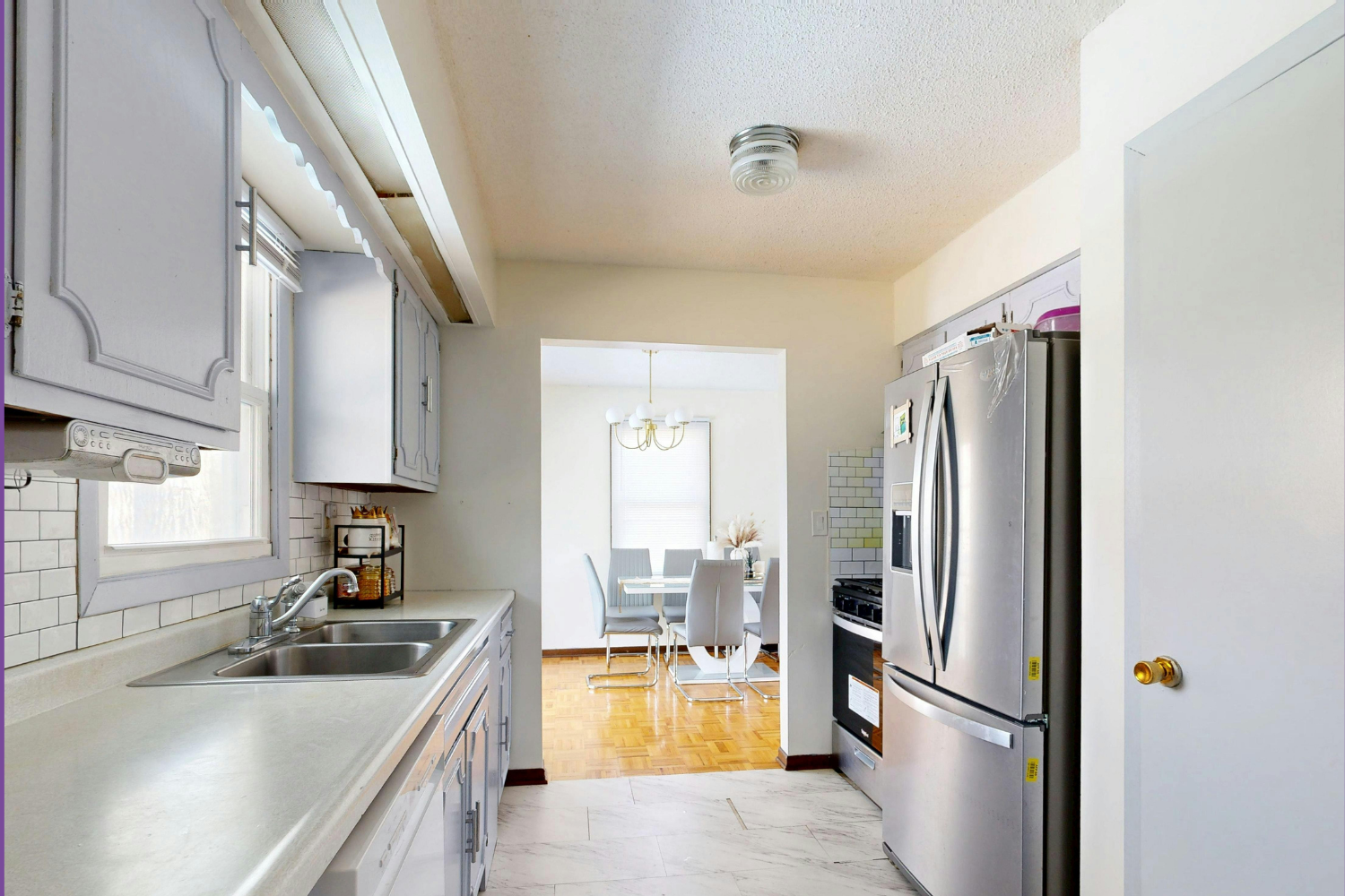 Interior view of a modest residential galley kitchen with light grey cabinets and white tile flooring. On the right is a stainless steel French-door refrigerator; on the left is a counter with a double sink under a window. An open doorway in the back leads to a dining area with a table and grey modern chairs.