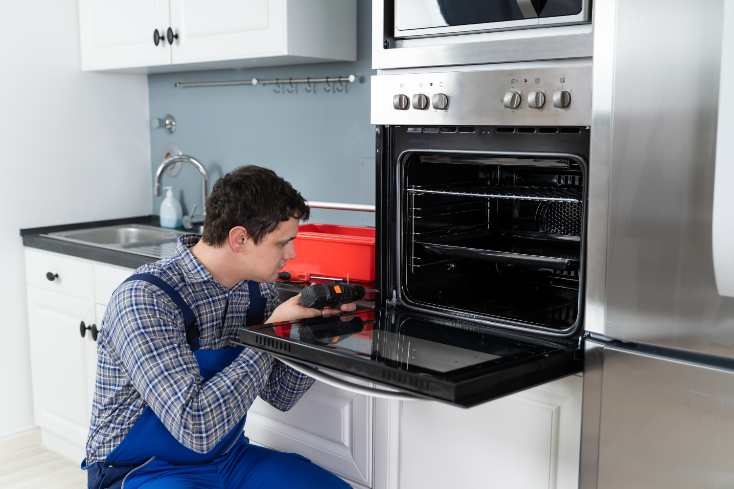 A technician repairs a stainless-steel oven with a red toolbox nearby.