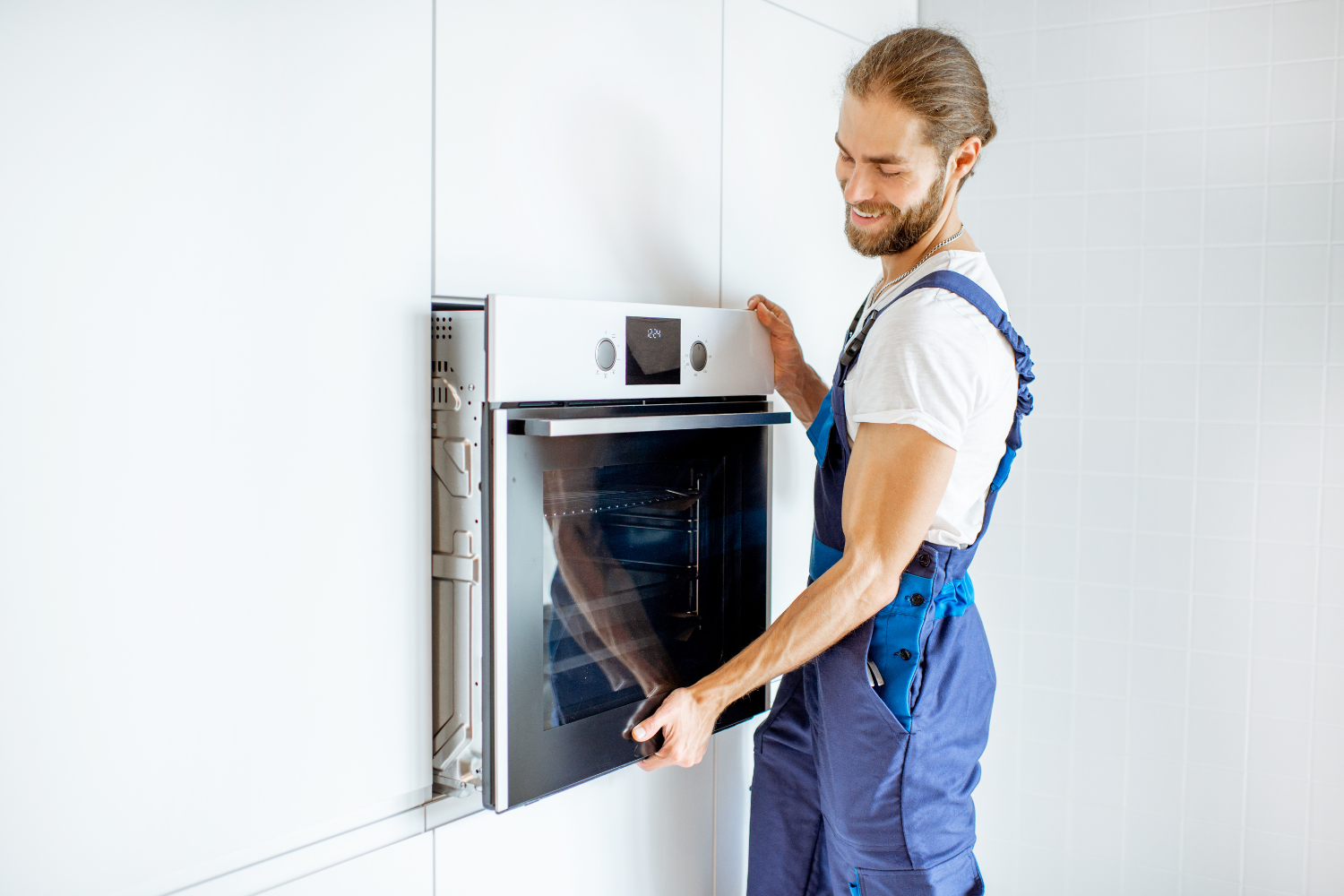 A person in blue overalls installs a built-in white oven into a white cabinet.