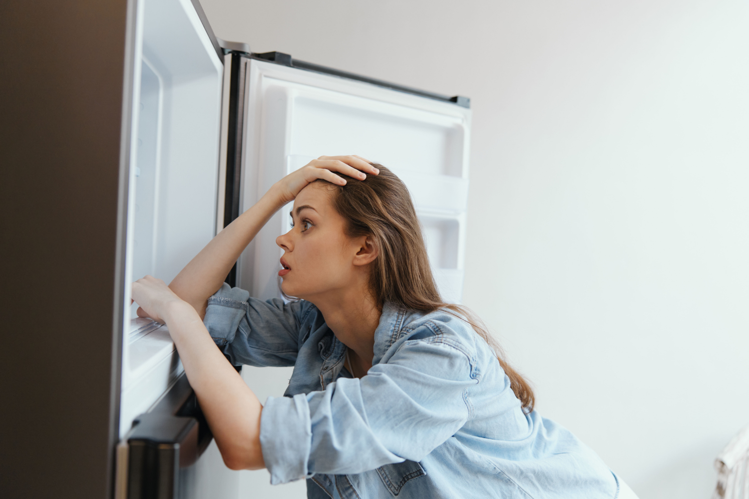 A person looking into an empty refrigerator with their hand on their forehead.