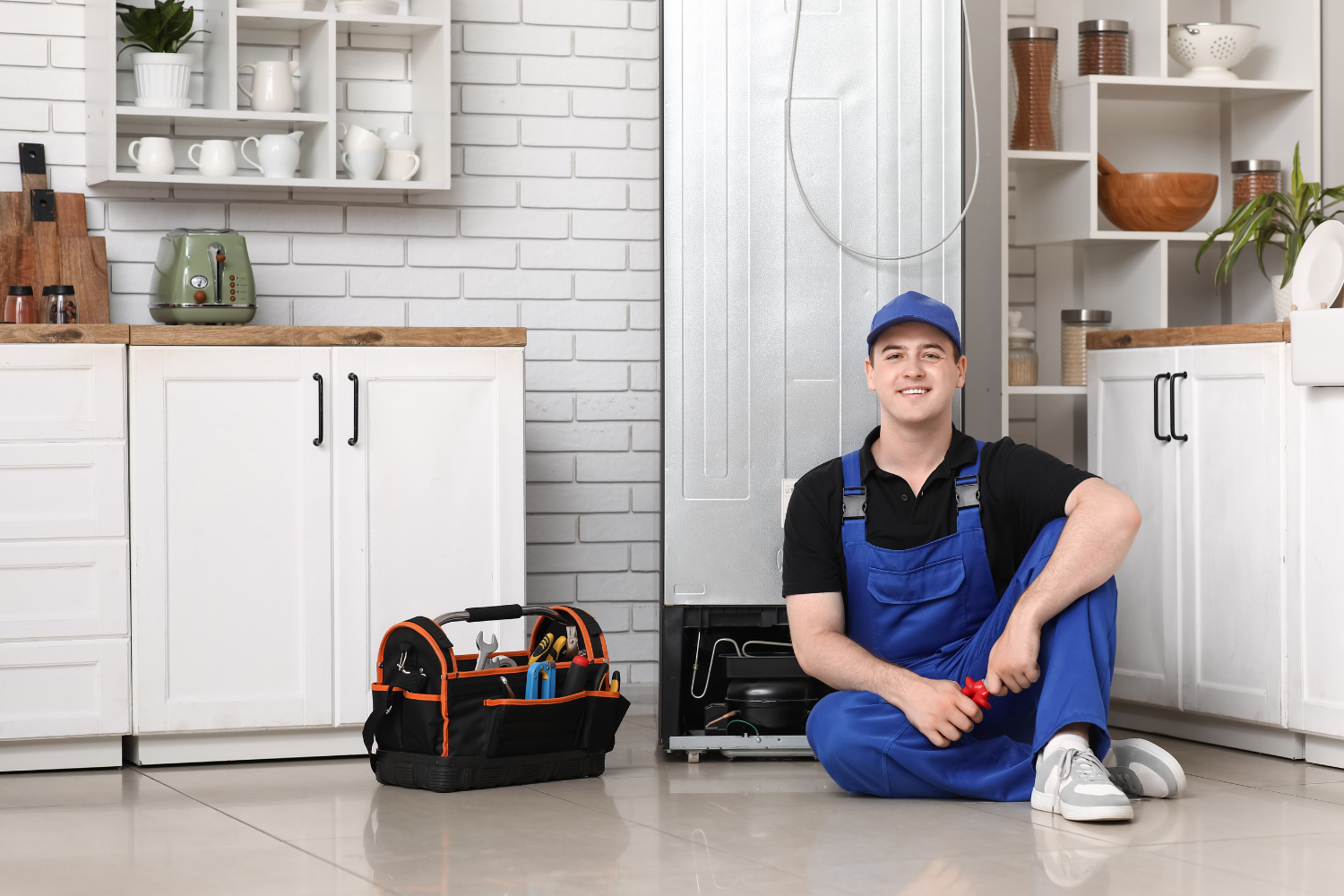 A repairman in blue overalls and a cap sits on the floor in a kitchen next to a toolbox and a refrigerator, smiling at the camera.