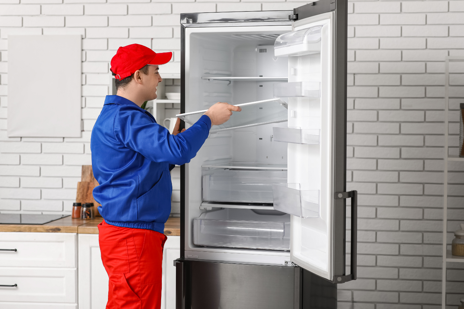 A person in a red cap and blue uniform is cleaning the inside of a refrigerator. The refrigerator door is open, and the person is holding a shelf with both hands.