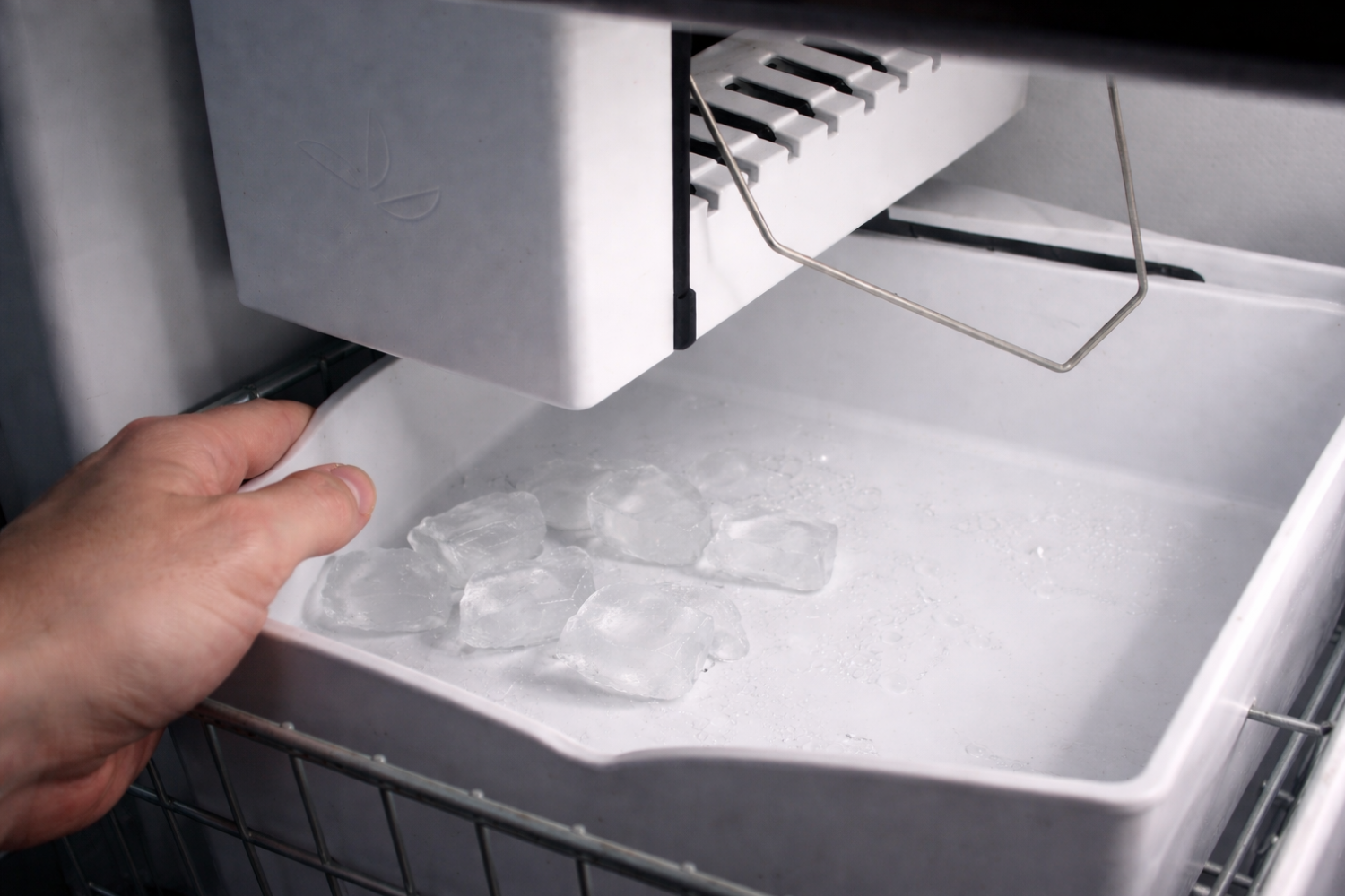 A hand is reaching into an ice maker to grab a white ice cube tray with only few ice cubes.