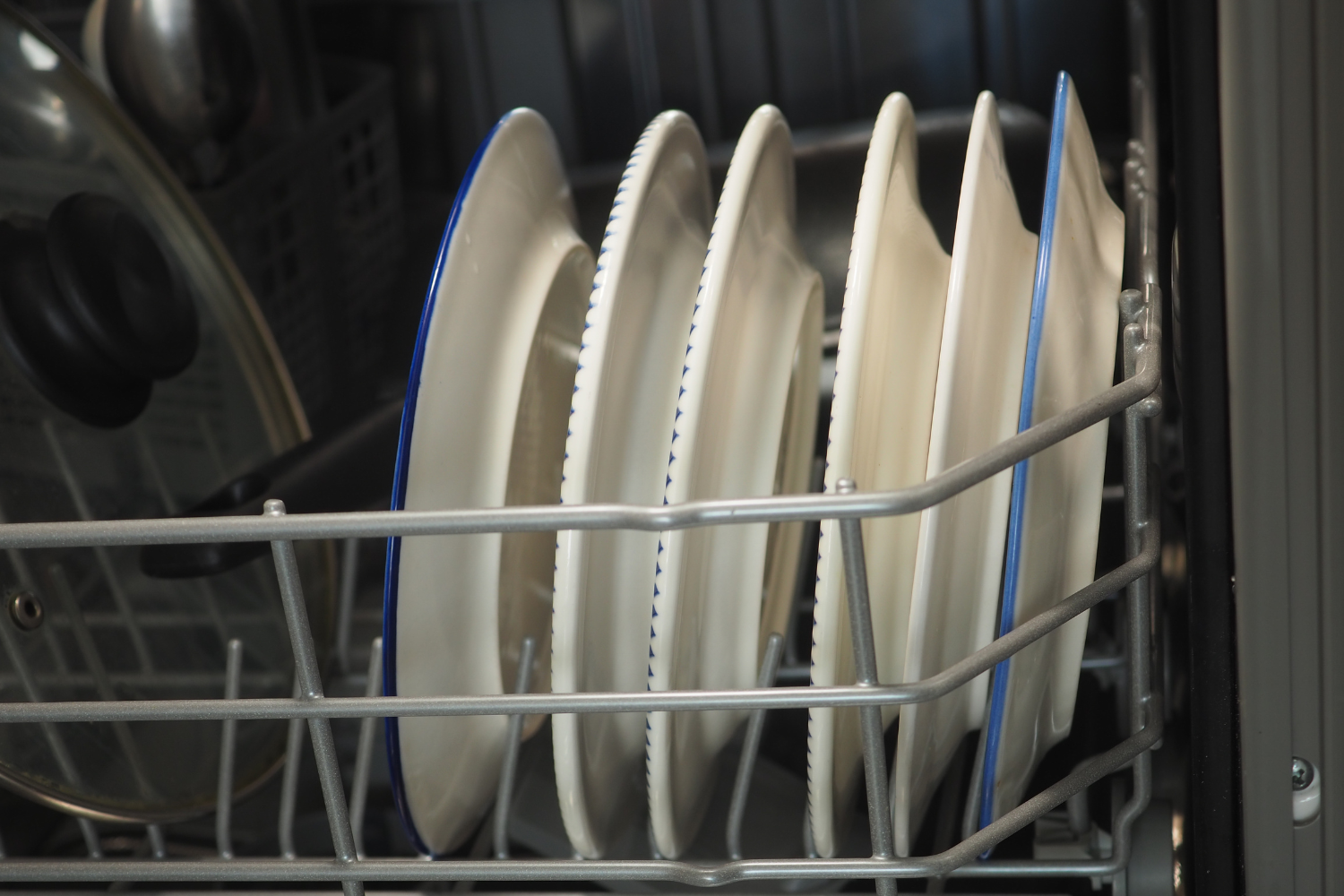 Several white plates with blue rims are arranged vertically in a dishwasher rack. A pot lid is visible to the left.