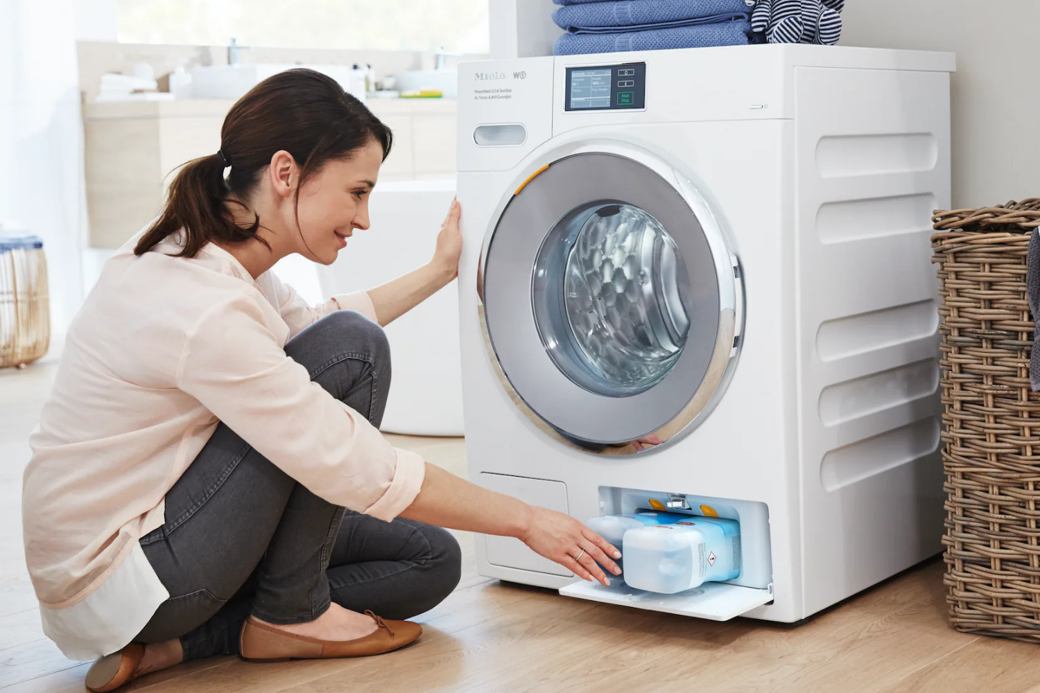 A person is shown tending to a washing machine, interacting with the detergent dispenser drawer. Next to the washing machine is a wicker laundry basket with towels draped over it.