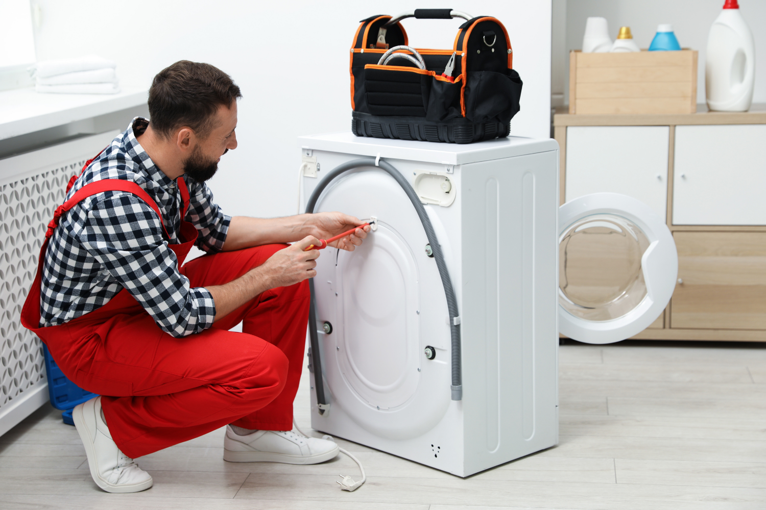 A person in red overalls and a checkered shirt kneels to repair a washing machine with a screwdriver, a toolbox rests on top.