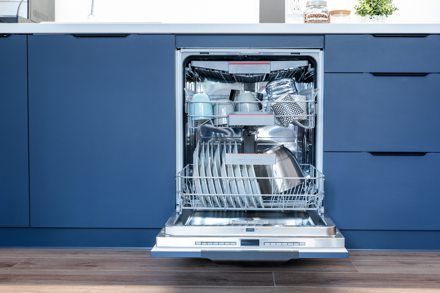 An open dishwasher filled with clean dishes, including plates, cups, and pots, set against a backdrop of blue kitchen cabinets.