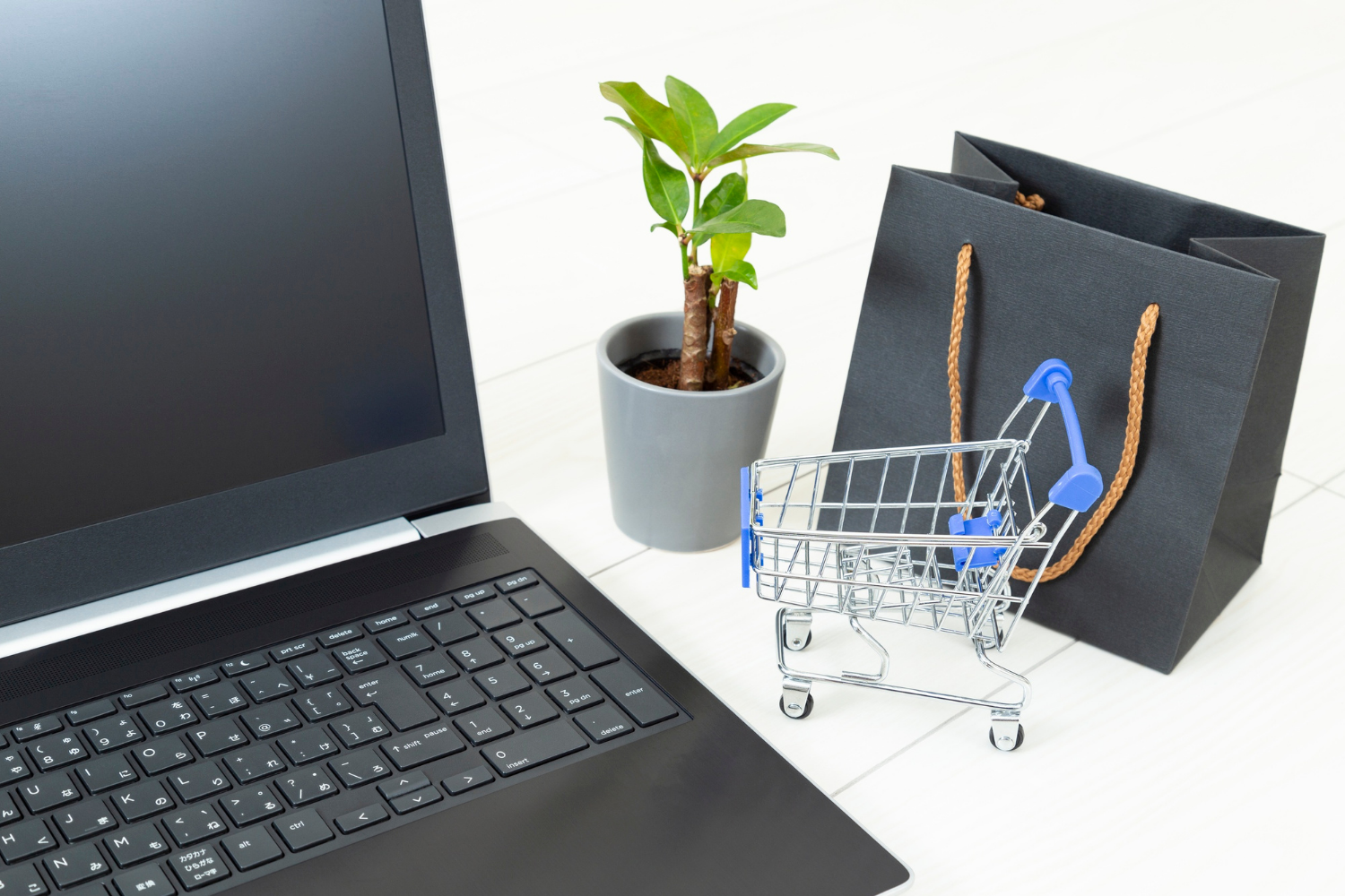 A laptop, a potted plant, a shopping bag, and a miniature shopping cart are arranged on a white surface, suggesting online shopping.