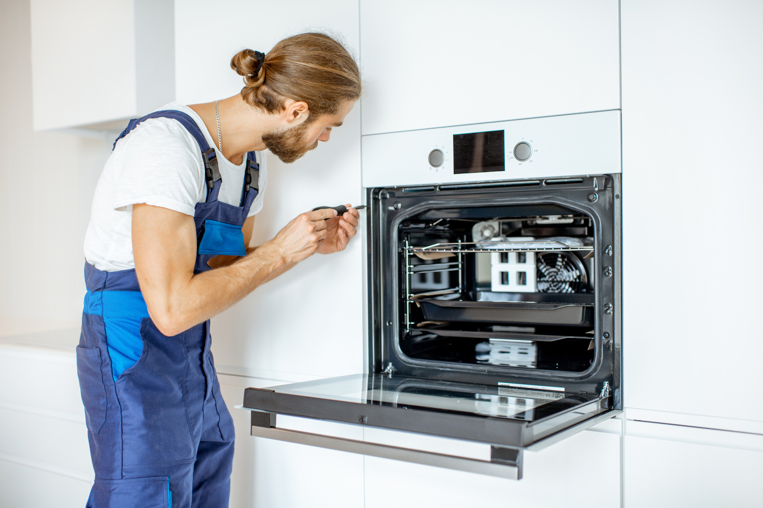 A person in blue overalls is fixing a built-in oven with a screwdriver.
