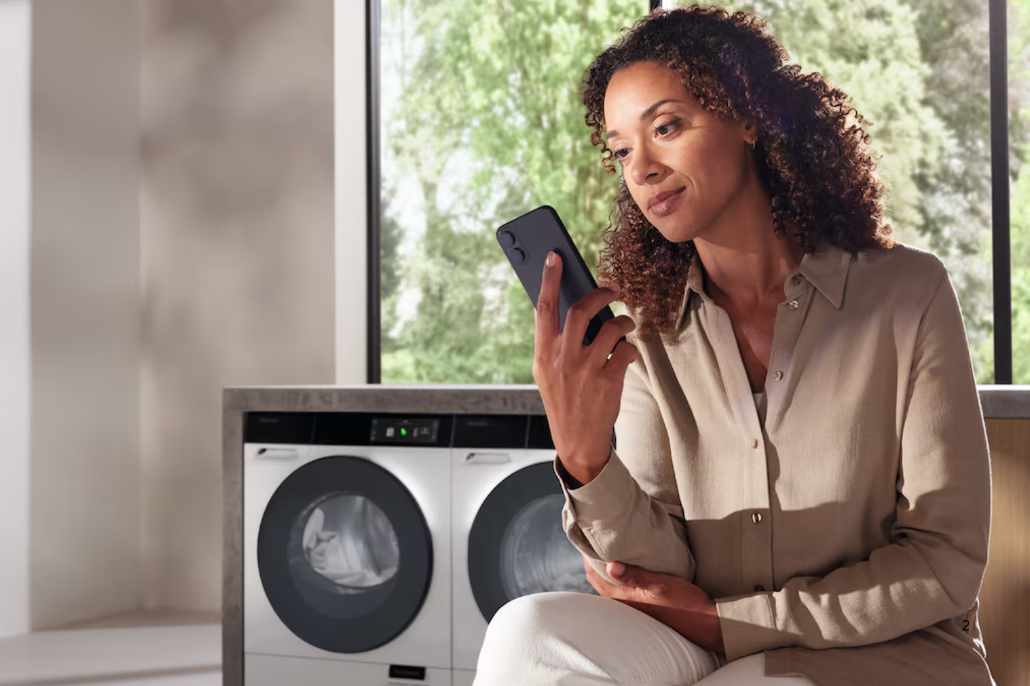 A person holds a smartphone while sitting in front of a stacked washer and dryer unit with a large window in the background.