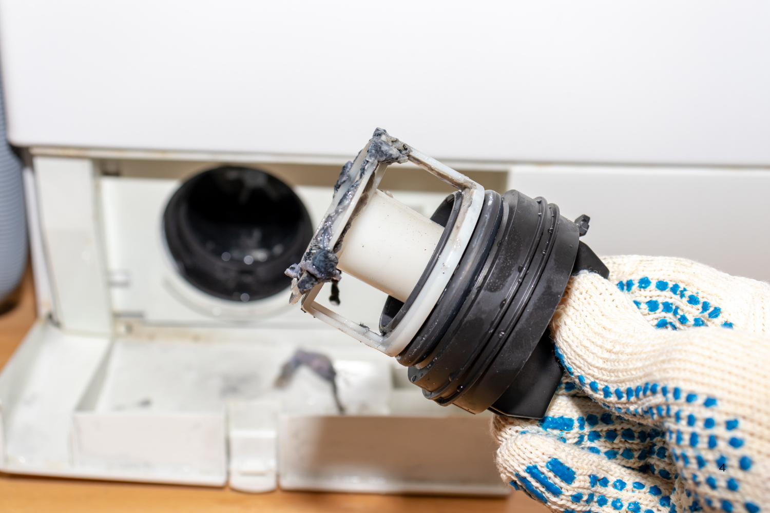 A gloved hand holds a dirty washing machine drain pump filter, with the open filter compartment visible in the background.
