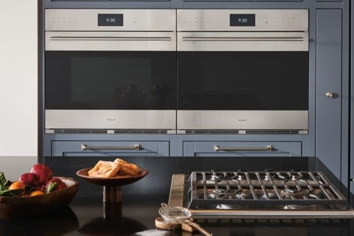 Two Wolf brand ovens are mounted above blue cabinets, with a gas stovetop in the foreground. A bowl of fruit and a dish of pastries sit on the counter next to the stovetop.
