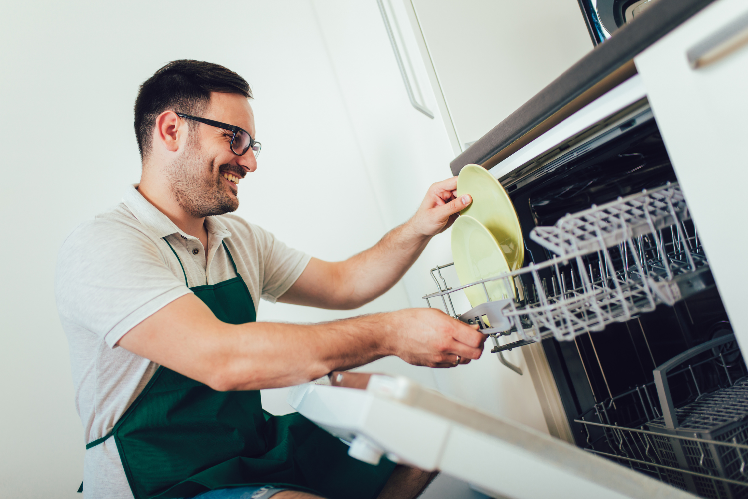 A person wearing an apron and glasses is loading light green plates into a dishwasher rack.