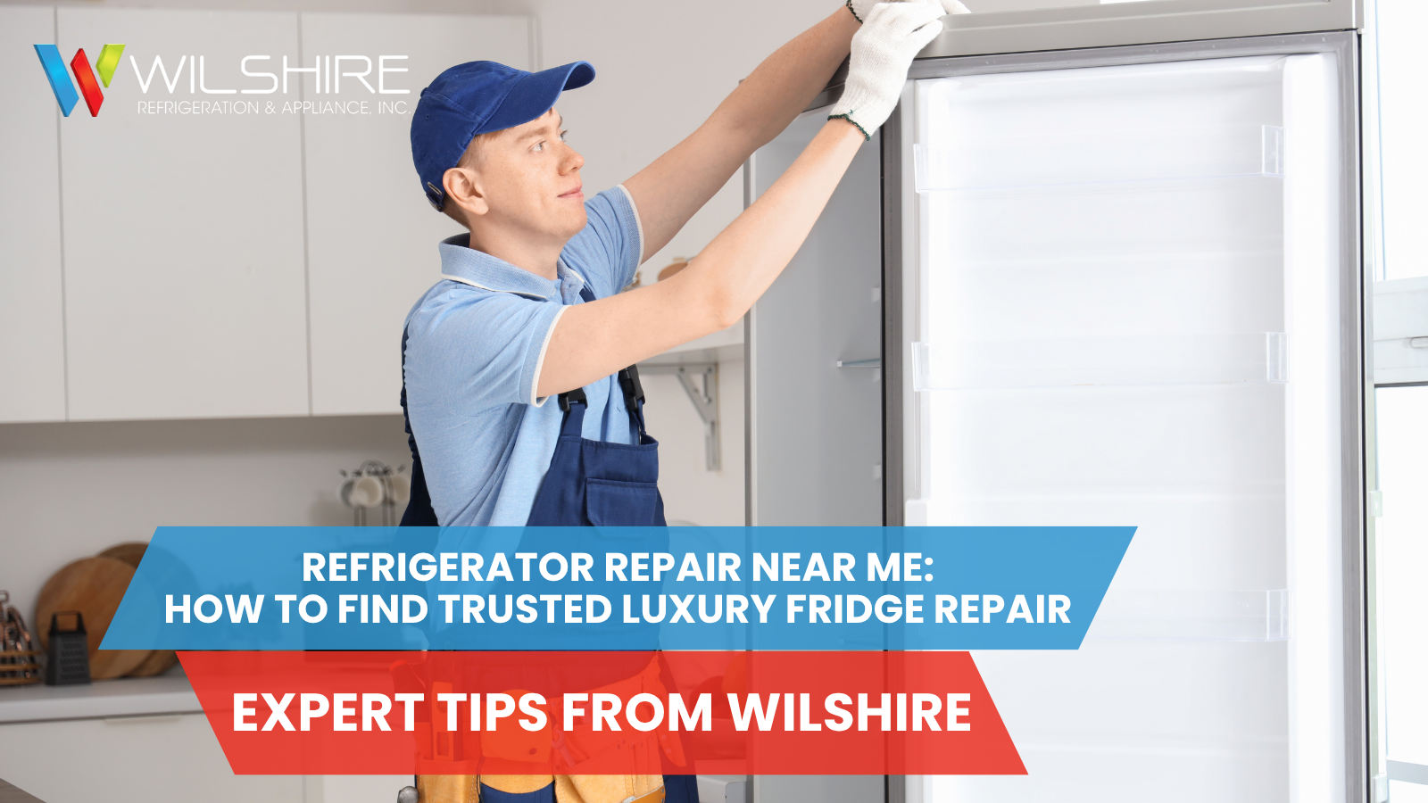 A repair person wearing a blue cap, blue shirt, and blue overalls with a tool belt is inspecting the top of an open refrigerator.