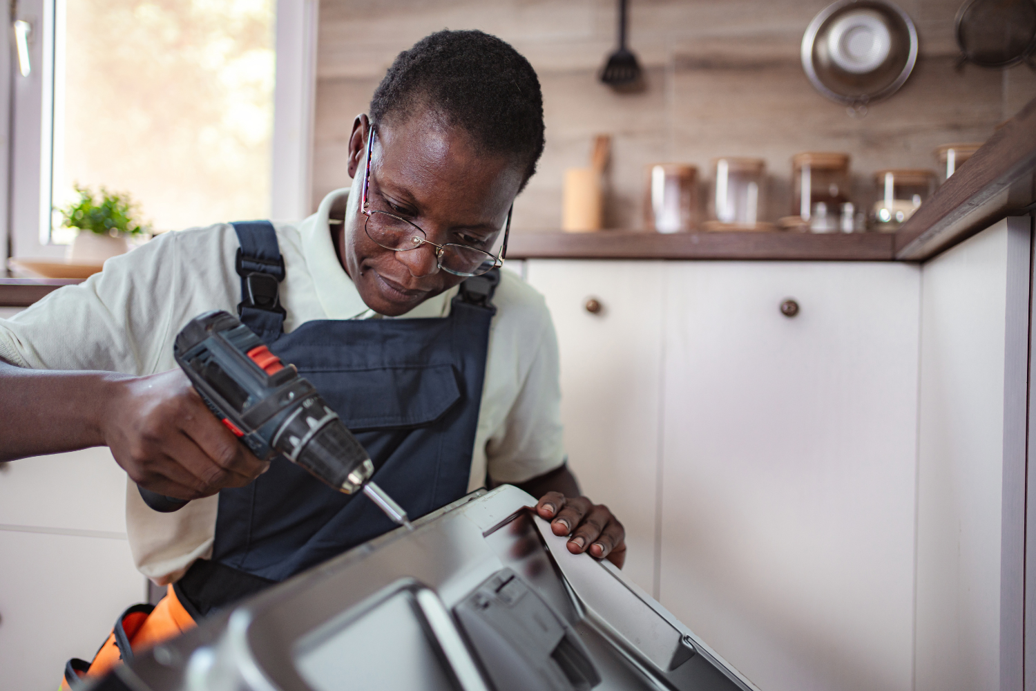 A person wearing overalls and glasses uses a cordless drill to screw into a metal appliance in a kitchen.