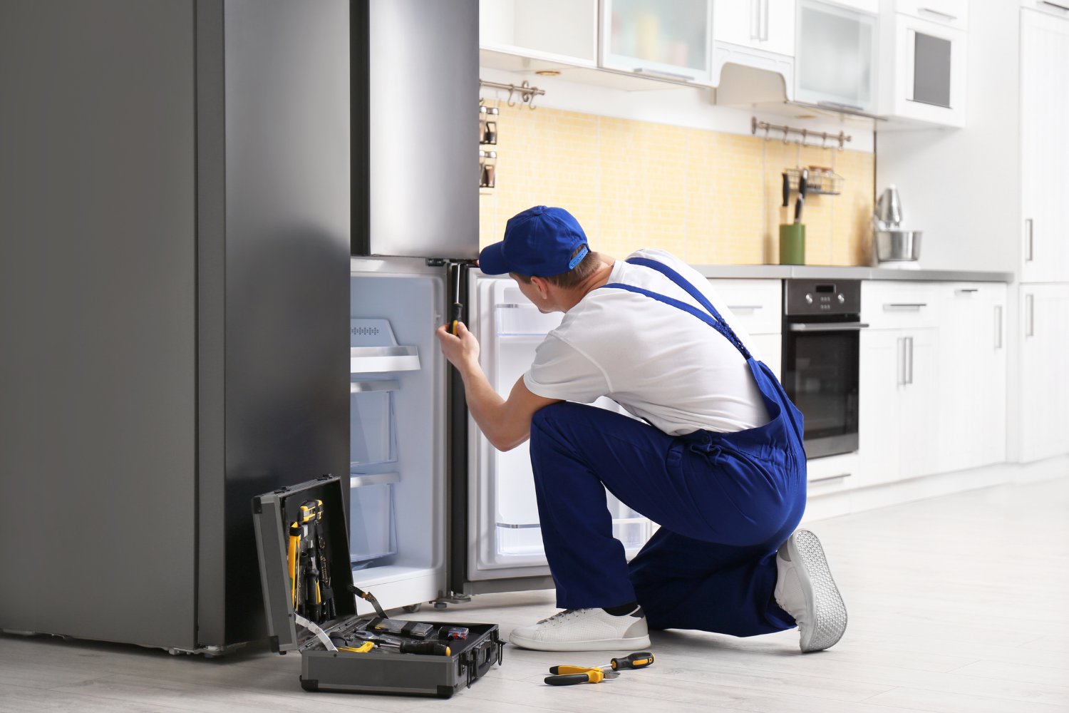 A technician in blue overalls and a cap kneels to repair an open refrigerator, with a toolbox and tools on the floor nearby.