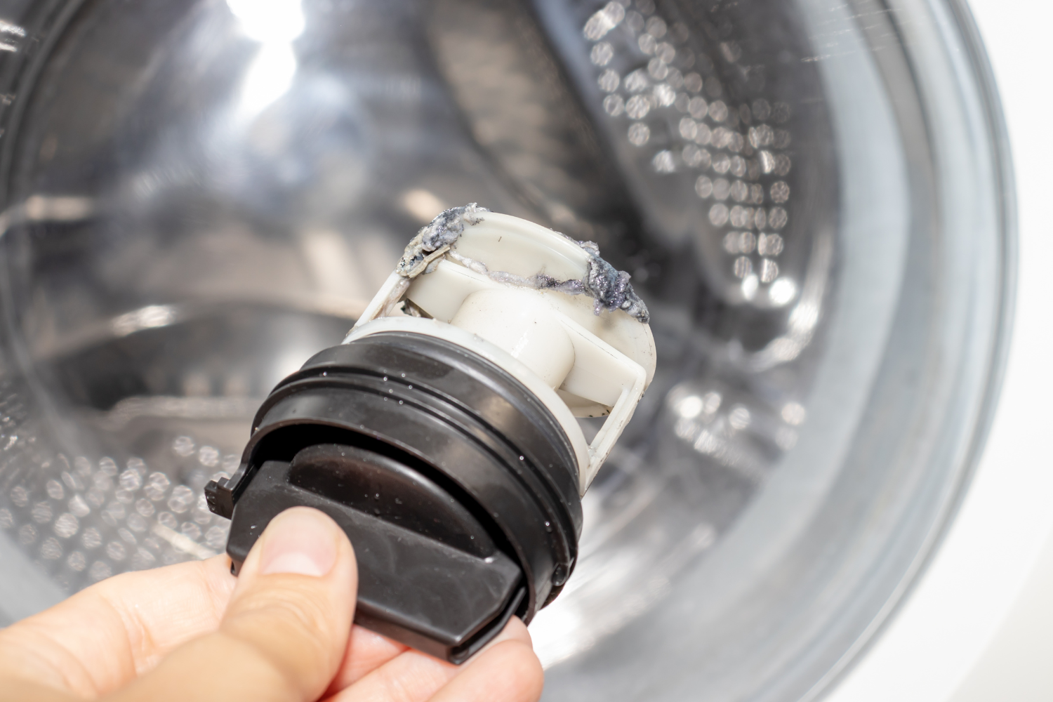 A close-up of a hand holding a plastic washing machine drain pump filter covered in gray, wet lint and grime, held up against the blurred background of a shiny, open washer drum.