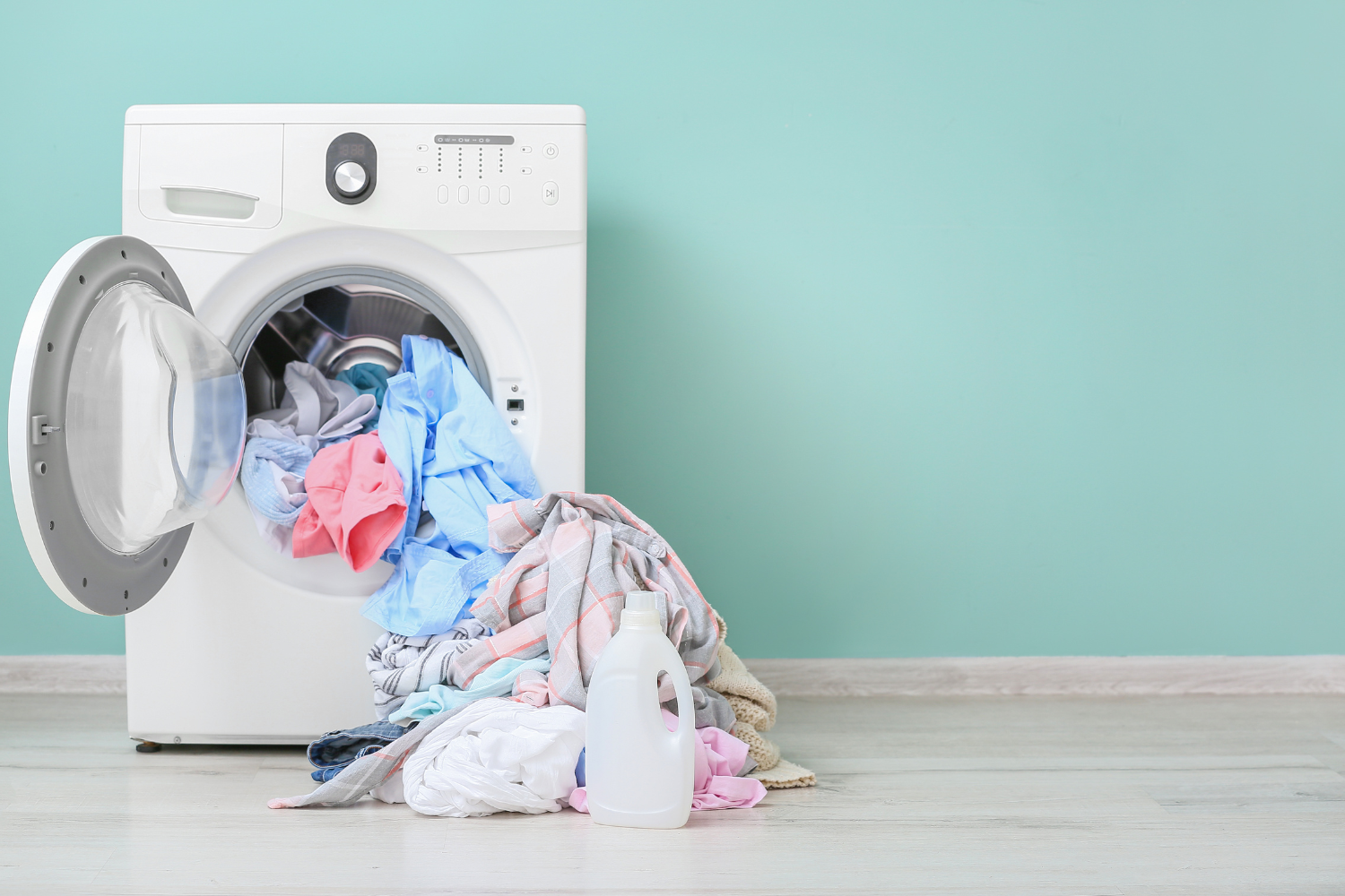 A washing machine with its door open and clothes spilling out onto the floor next to a bottle of detergent.