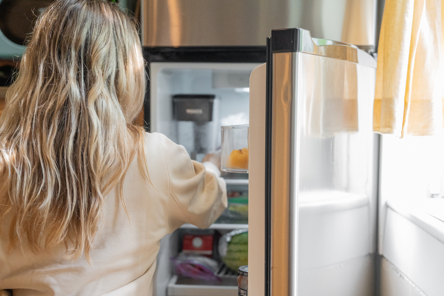 A person with blonde hair reaches into an open refrigerator, holding a clear container with an orange item inside.