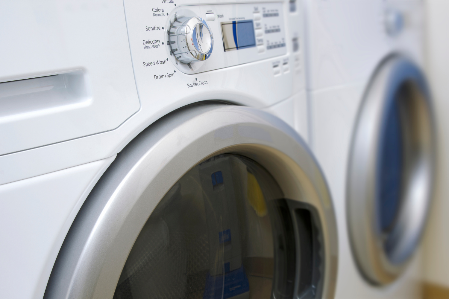 Two individuals install new washing machines in a laundry room, with one person leaning over a machine and the other wearing gloves.