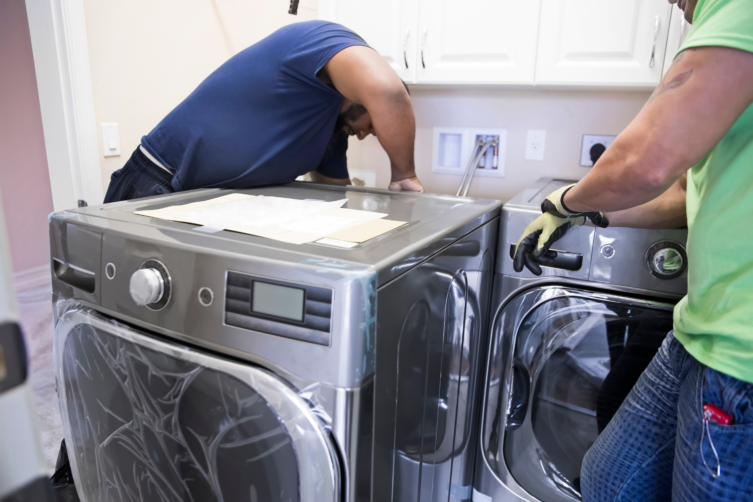 Two individuals install new washing machines in a laundry room, with one person leaning over a machine and the other wearing gloves.