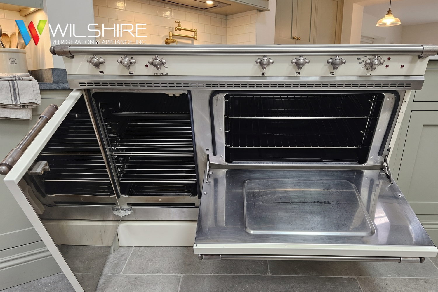 Commercial-style stainless Lacanche steel oven with the front door open, showing multiple black oven racks and a warming drawer, set in a light-colored kitchen with pendant lights and tiled flooring.