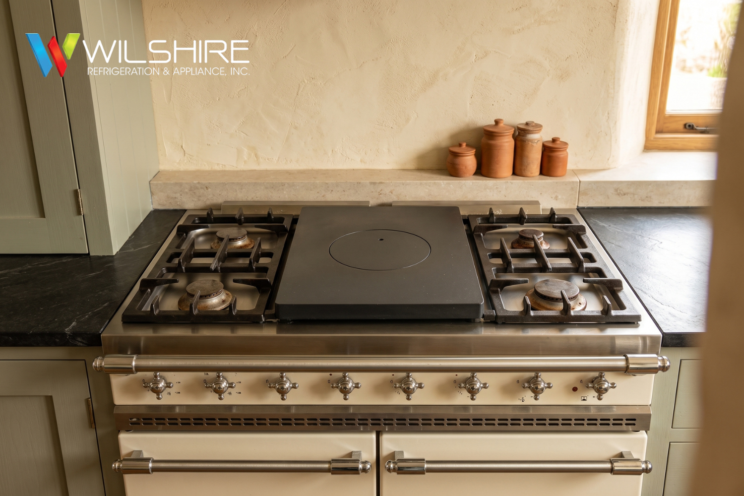 Built-in stainless steel Lacanche gas range with two side burners, a central black glass cooktop with a round burner outline, and storage drawers beneath, set in a light stone surround by a window.
