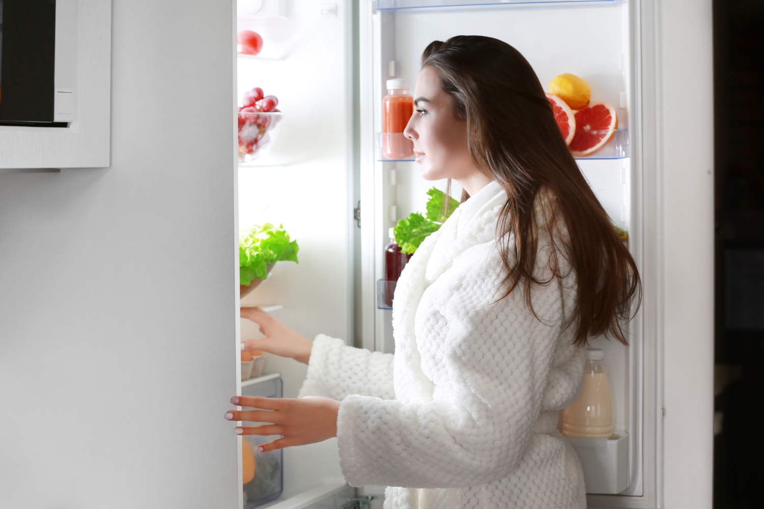A person in a white bathrobe opens a refrigerator door, reaching toward shelves with jars, greens, and fruit visible inside.