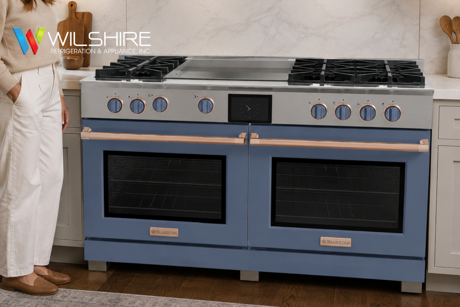 A person in a light sweater and white pants stands beside a blue-and-white freestanding Bluestar kitchen range with four burners, two ovens, and control knobs in a bright modern kitchen.