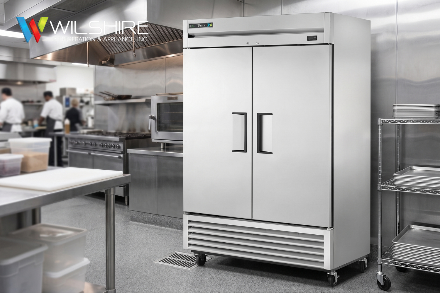 Stainless-steel commercial kitchen with prep tables and ovens, and a large double-door white True refrigeration unit on wheels in the foreground.